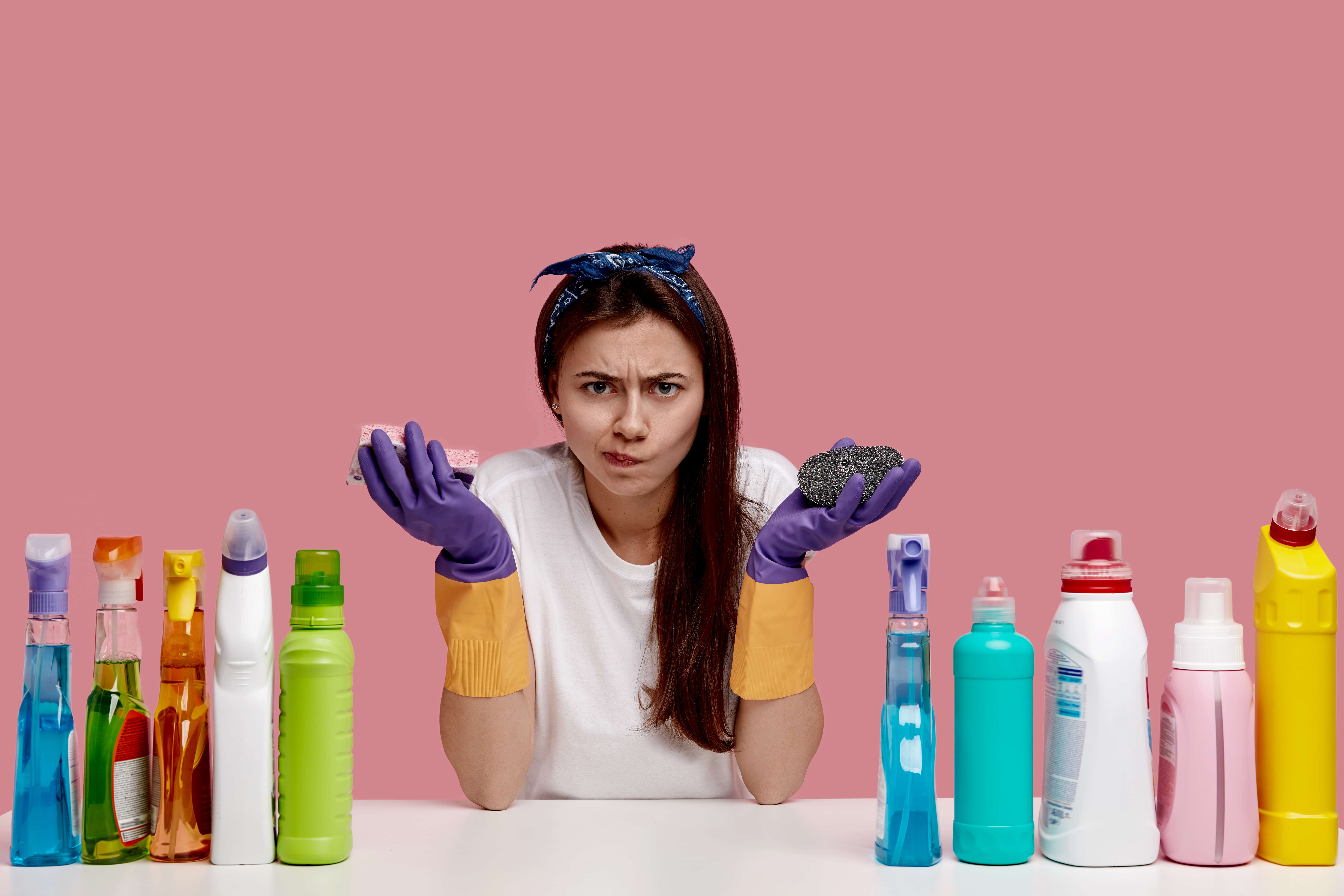 Displeased young woman frowns face in discontent, carries sponges, wears white casual t shirt, rubber gloves, looks angrily in camera, uses different cleaning supplies, isolated over pink background